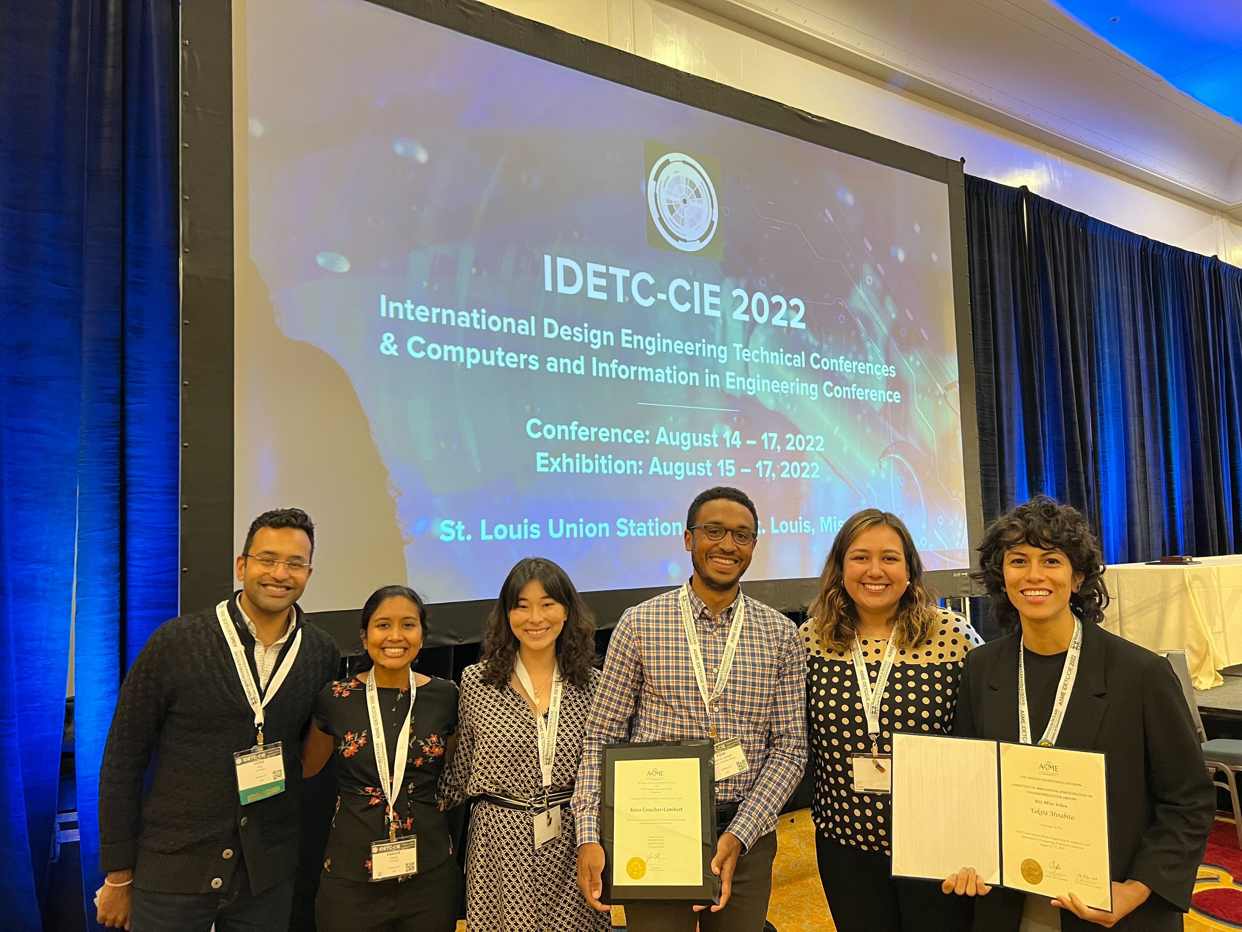 from left to right, Vivek, me (Ananya), Elisa, Kosa (with award plaque), Nicole, and Yakira (with certificate) in front of projector screen with the IDETC conference name on it