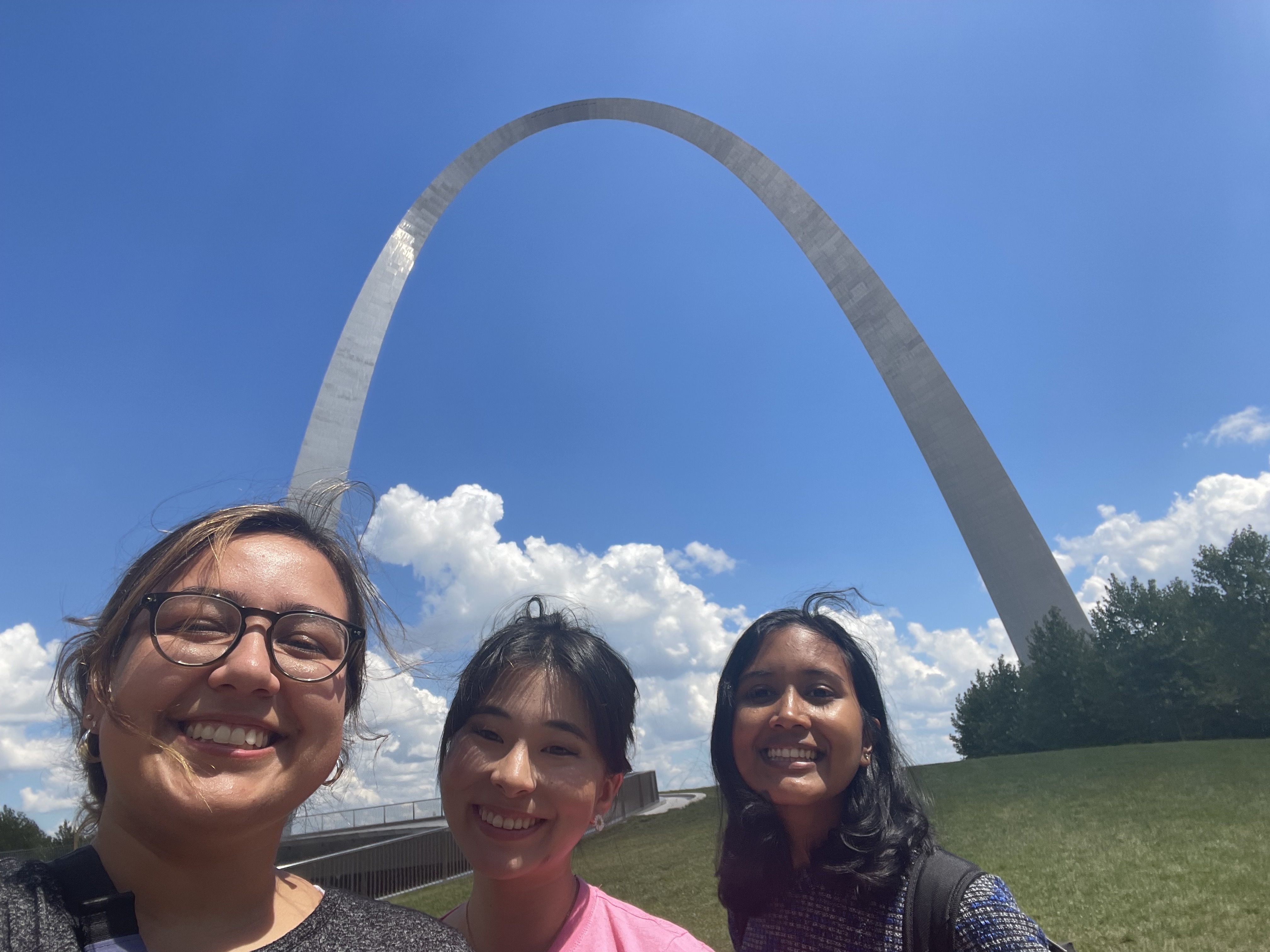 from left to right, selfie of Nicole, Elisa, and me (Ananya) in front of the metal St. Louis Gateway Arch with blue sky and clouds in the background